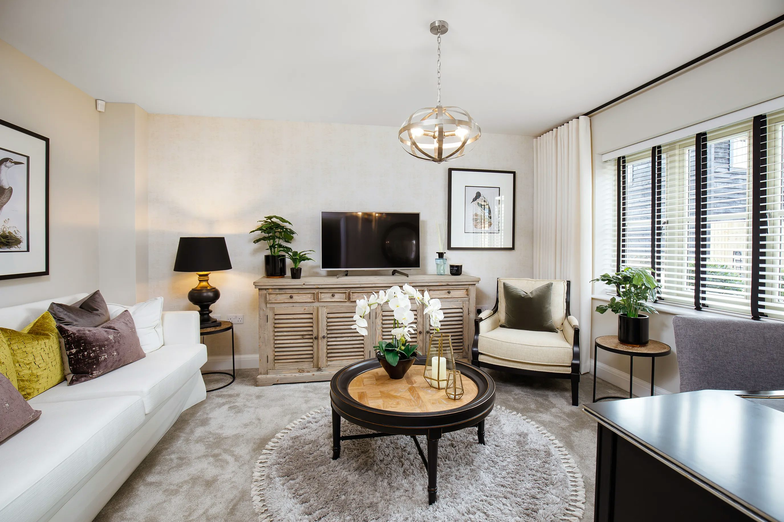 A modern living room with a white sofa adorned with colorful cushions, a round wooden coffee table with a potted plant and candles, and a light-colored armchair. A wooden sideboard sits beneath a wall-mounted flat-screen TV, accompanied by framed pictures, a black table lamp, and additional potted plants. The space features a neutral color palette accented with green and gold, illuminated by natural light filtering through window blinds. A modern chandelier hangs from the ceiling, complementing the contemporary interior design.
