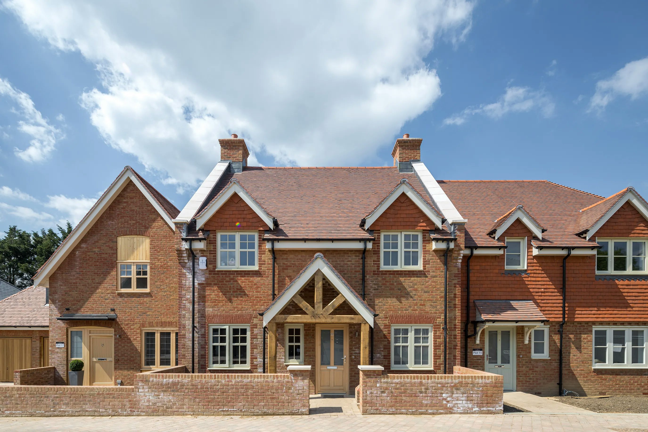 A row of newly built, modern brick houses with pitched roofs and chimneys. The central house features a wooden porch with double doors, adding a welcoming element. The houses have multiple windows, arranged symmetrically to enhance the architectural balance. Above, the sky is mostly clear with a few scattered clouds, suggesting pleasant weather.