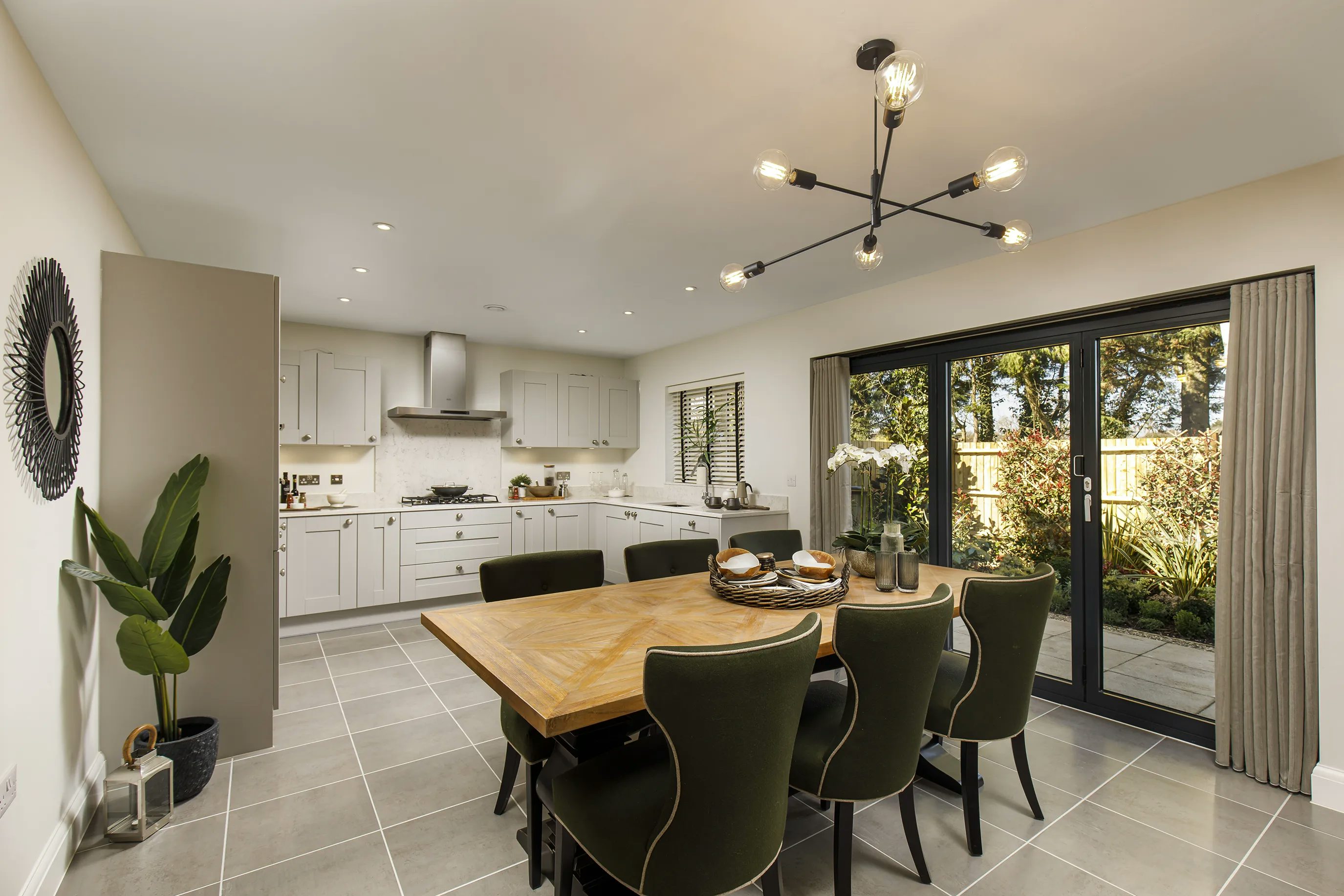 A modern kitchen and dining area with white cabinetry, a stainless steel range hood, and various kitchen appliances. A wooden table with six green upholstered chairs sits under a contemporary chandelier with exposed bulbs. Large glass doors open to an outdoor patio with greenery and a wooden fence, allowing in natural light. Decorative elements include a wall mirror and potted plants.