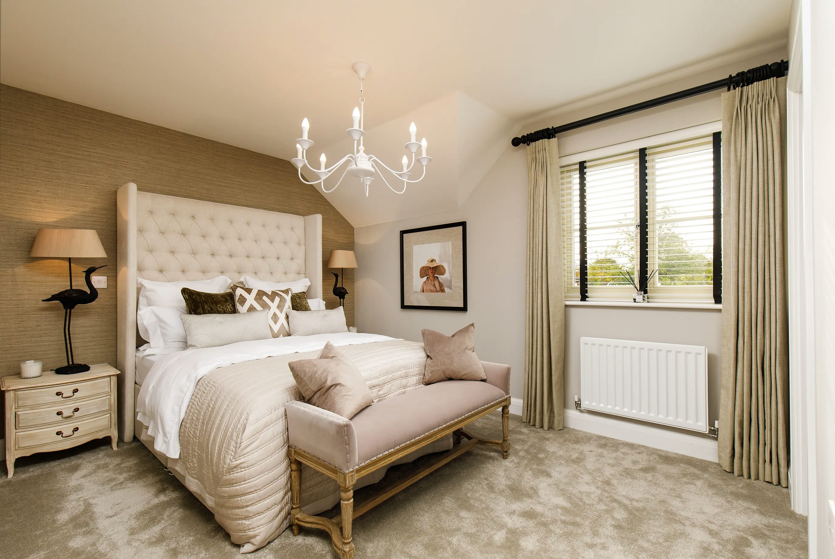 A well-decorated bedroom with a beige and neutral colour scheme. The room features a large bed with a tufted headboard, adorned with multiple pillows and a beige comforter. A cushioned bench sits at the foot of the bed. On either side, matching nightstands hold lamps. The wall behind the bed has textured wallpaper, with framed artwork above. A chandelier hangs from the ceiling, casting elegant lighting. A window with beige curtains and blinds sits above a white radiator. The carpet is light beige, complementing the overall aesthetic.
