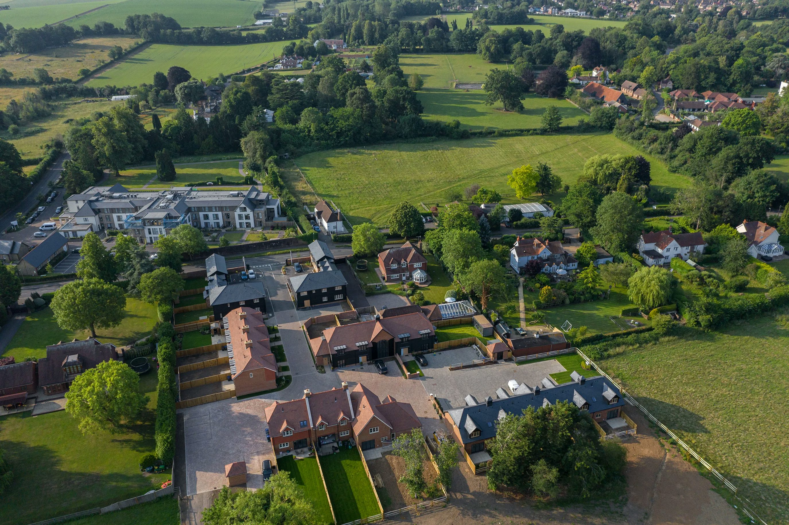 Aerial view of a residential neighborhood with houses arranged in a structured layout, surrounded by green fields and trees. The homes feature well-maintained gardens and driveways, with additional houses and open land visible in the distance.