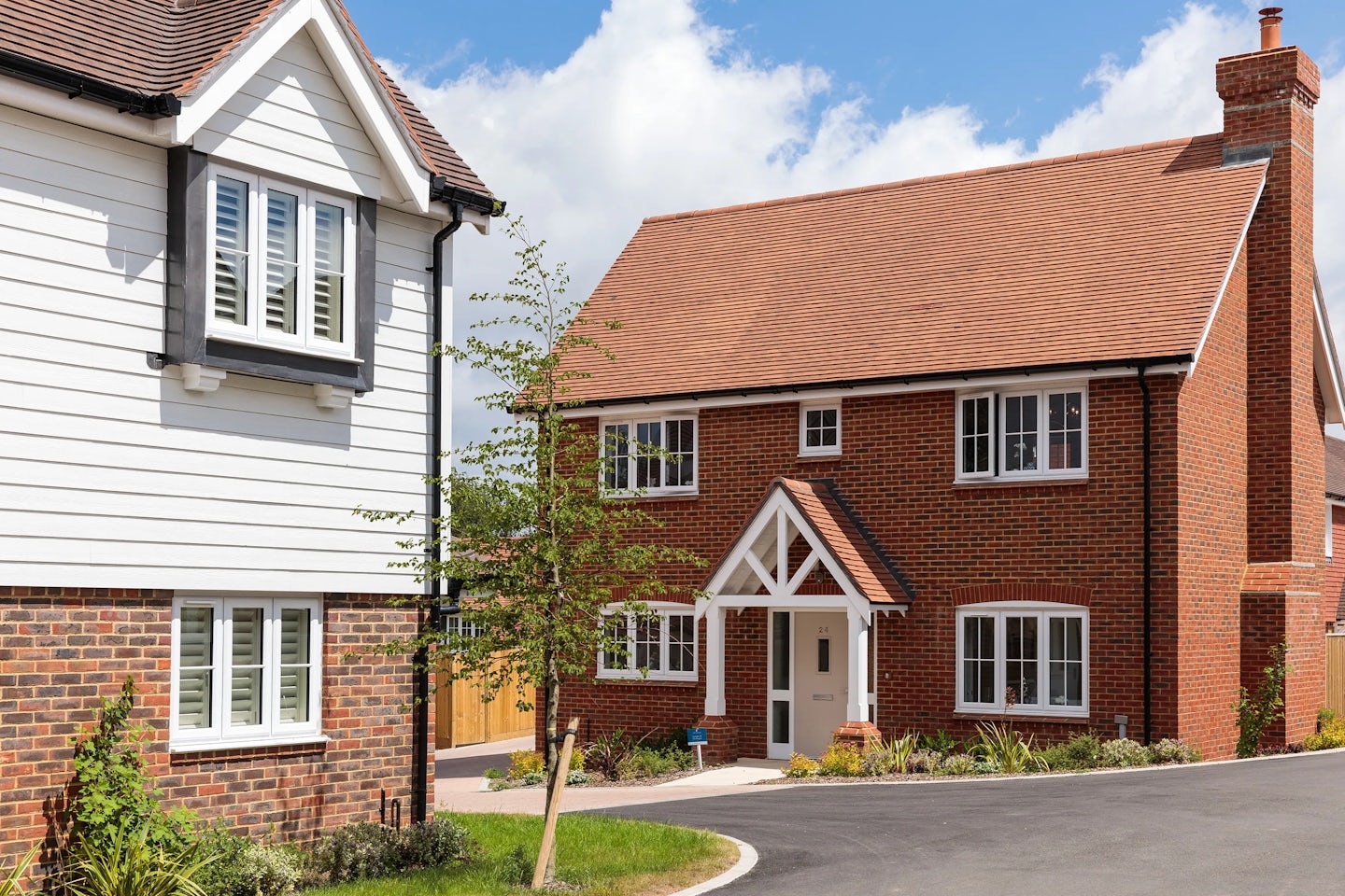 A two-story brick house with a gabled roof, white-framed windows, and a white front door with a small porch. To the left, a partially visible house with white siding and black window trim. A small tree and landscaping in front. The sky is blue with scattered clouds.