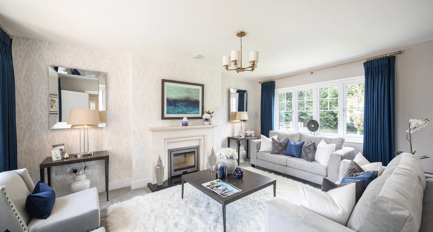 A modern living room with elegant decor. The space features a large window with blue curtains, allowing natural light to enter. Two sofas, adorned with pillows in shades of blue, white, and gray, face each other. A coffee table sits in the center, holding decorative items and magazines. A fireplace with a painting above it is flanked by two side tables with lamps. A chandelier hangs from the ceiling, complementing the sophisticated ambiance. The walls have patterned wallpaper, and a large mirror is mounted on the left wall.
