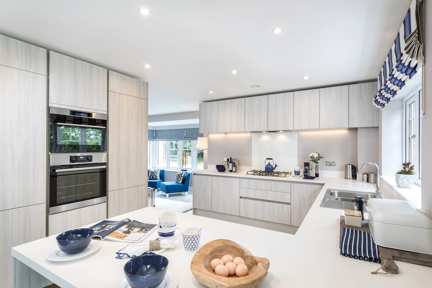 A modern kitchen with light wood cabinetry and white countertops. Built-in appliances include an oven, microwave, and gas stove with a range hood. A sink with a faucet is positioned near a window with blue and white striped blinds. Various kitchen items, such as a bowl of eggs, blue bowls, a newspaper, and a cup, are placed on the countertops. Recessed lighting illuminates the space. In the background, a living area with blue chairs and a large window is partially visible.