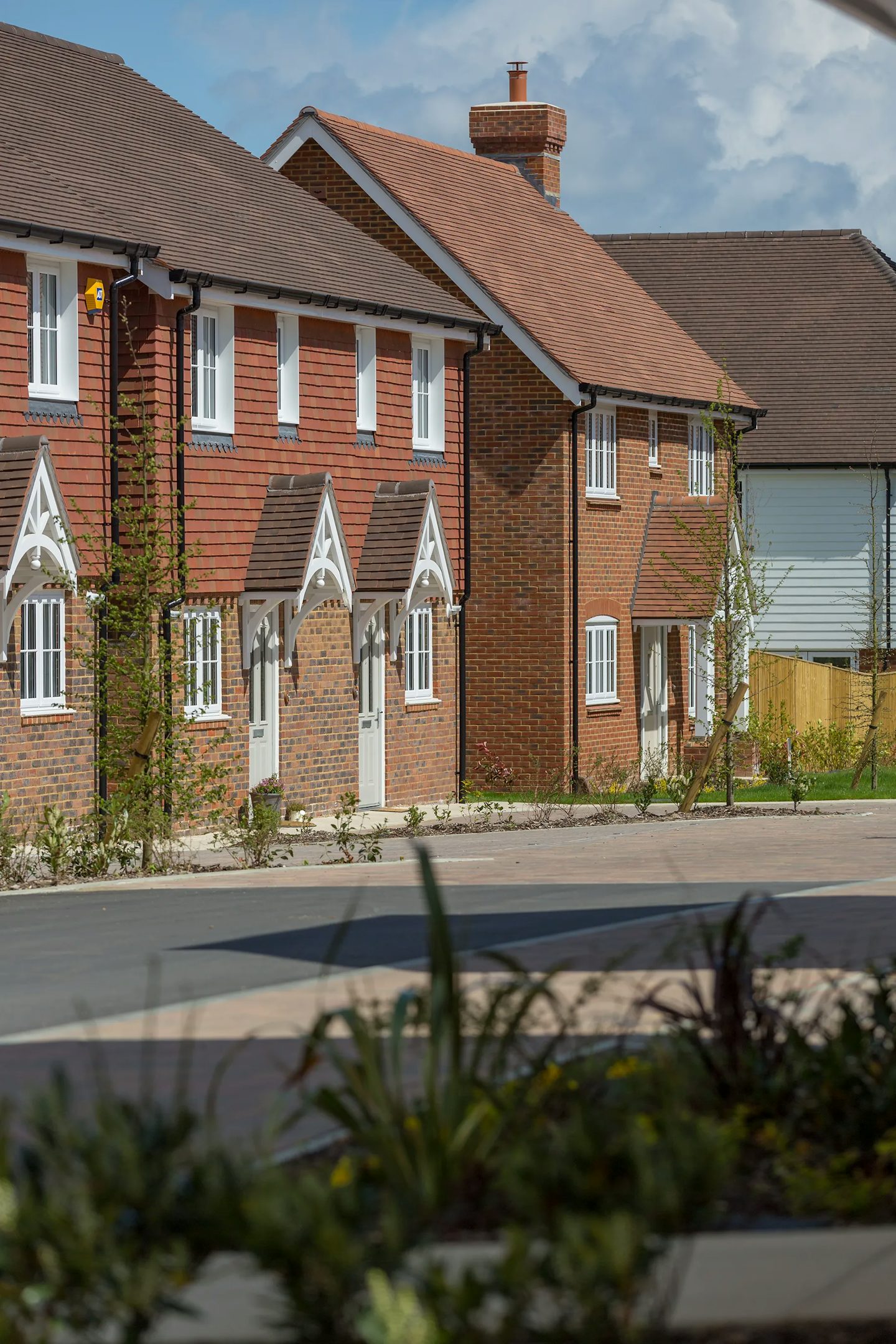 A row of modern suburban houses with brick and wooden facades, pitched roofs, and white-framed windows and doors. A well-maintained garden with various plants and shrubs is in the foreground. The scene is bright and sunny, suggesting a pleasant neighbourhood.