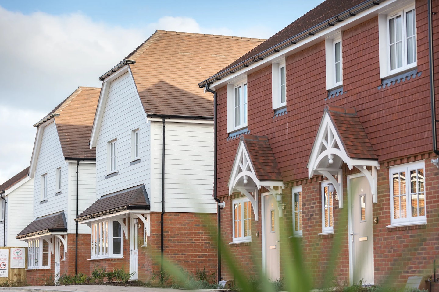 A row of modern suburban houses with white and red brick facades, pitched roofs, and small front porches. The architecture blends traditional and contemporary elements. A grassy area is in the foreground, and the sky is partly cloudy.