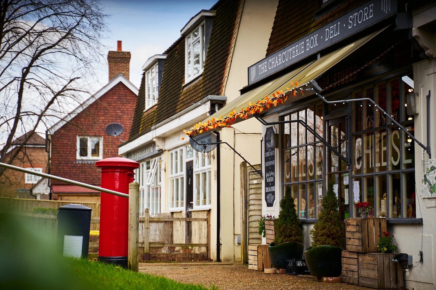 A traditional street scene featuring "The Charcuterie Box - Deli - Store," a shop with an autumn-themed awning and a window displaying a large "CHEESE" sign. A red postbox and black trash bin stand in the foreground. The background includes a brick house with white-framed windows and a satellite dish, with bare trees suggesting autumn or winter. Small potted plants are placed outside the shop entrance.