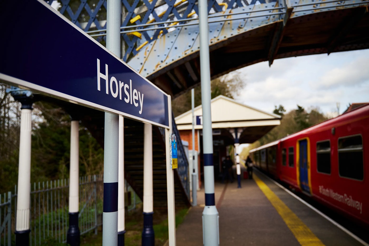 A South Western Railway train is stopped at Horsley station, positioned on the right side of the image. The platform has a yellow safety line along its edge. A pedestrian bridge spans across the tracks. In the background, trees and greenery frame the station, indicating a rural or suburban setting. The station sign displaying "Horsley" is prominently placed on the platform.