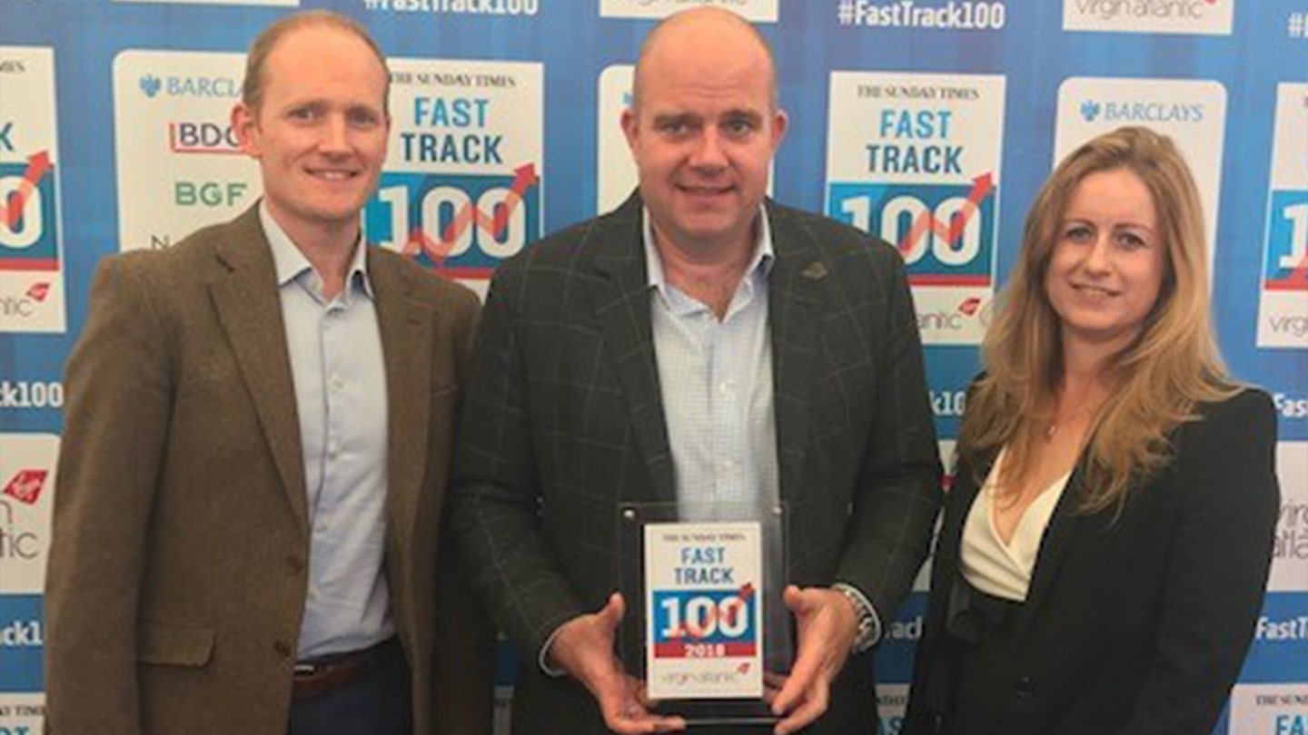 An award ceremony backdrop with logos from Barclays, BGF, and other sponsors, featuring the text "#FASTTRACK100" and "THE SUNDAY TIMES FAST TRACK 100." Three individuals stand in front of it, with the central person holding a plaque that reads "THE SUNDAY TIMES FAST TRACK 100 2018 virgin atlantic." The scene highlights a recognition event for the UK's fastest-growing companies.