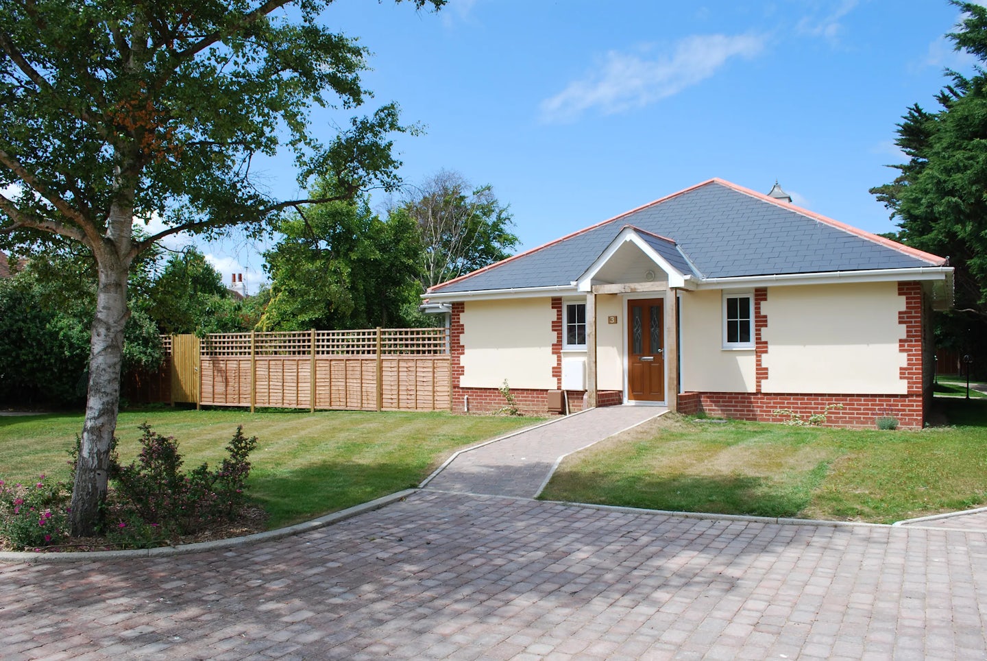 A single-story house with a grey tiled roof and beige exterior walls accented by red brickwork. The entrance features a wooden door with glass panels, flanked by two small windows. A paved path leads to the door, bordered by a neatly trimmed lawn and a garden. A wooden privacy fence lines the left side of the property. The sky above is clear and blue, suggesting a sunny day.