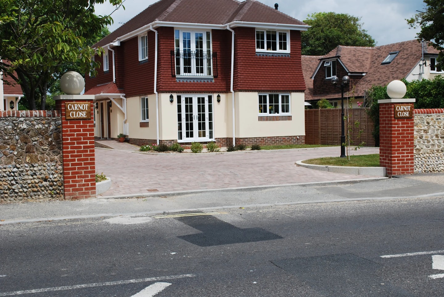 A two-story house with a red-tiled roof and upper red siding sits behind a low stone and brick wall. The lower half of the house exterior is cream-colored, with large windows and French doors on the ground floor, and a balcony above. The paved driveway, made of light bricks, is flanked by two brick pillars topped with spherical lights and signs that read “CARNOT CLOSE.” Trees and additional houses appear in the background. The setting is a tidy, suburban residential area.