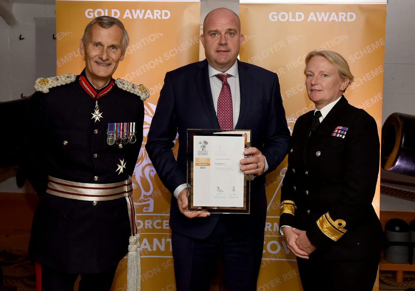 A man in a suit stands between two uniformed military personnel, holding a framed certificate. The backdrop behind them displays the words "GOLD AWARD" and "EMPLOYER RECOGNITION SCHEME". The military personnel, dressed in formal uniforms adorned with medals and insignia, flank the recipient at what appears to be an award ceremony recognizing employer support for the armed forces.