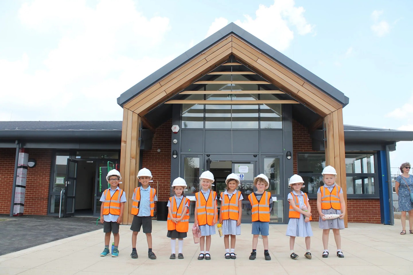 A group of eight children wearing orange safety vests and white construction helmets stand in front of a modern building. The building features a wooden frame at the entrance and large glass windows.