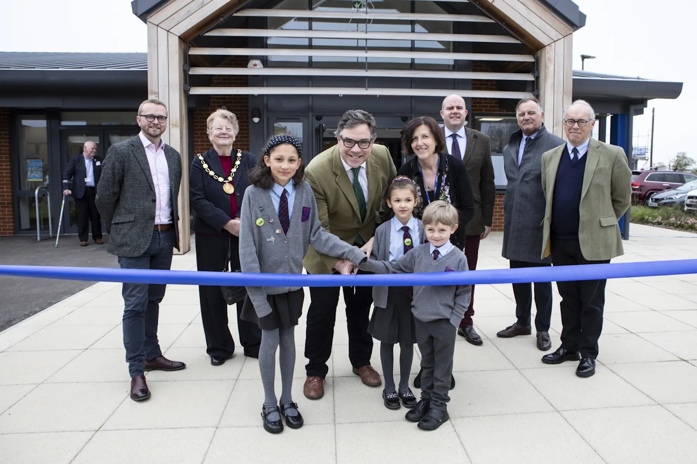 A group of nine people, including three children in school uniforms and six formally dressed adults, participate in a ribbon-cutting ceremony in front of a modern school building with a wooden frame and large glass windows. The children hold the ribbon while an adult in the centre uses scissors to cut it.