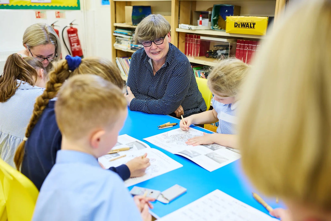 A group of children is seated around a blue table, working on worksheets. An adult is sitting with them, overseeing their work. The background includes shelves with books, a yellow box labeled "DEWALT," and two fire extinguishers mounted on the wall. The children appear engaged in a classroom activity, possibly solving math problems. The worksheets contain arithmetic exercises such as addition, subtraction, multiplication, and division.