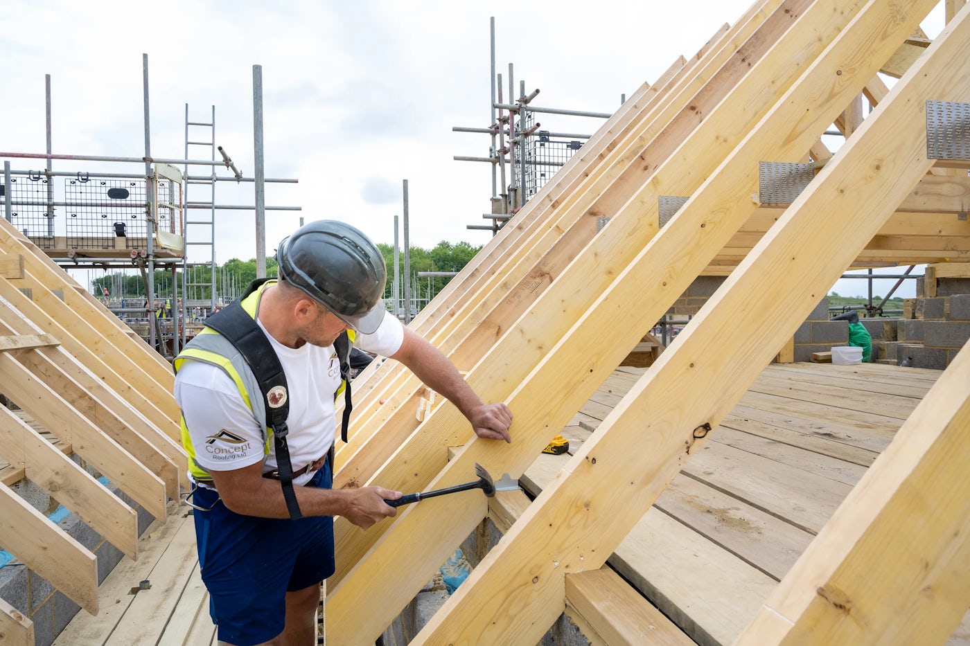 A construction worker in a hard hat and safety vest is hammering a wooden beam at an outdoor construction site. The structure appears to be a roof frame, with multiple wooden beams and scaffolding visible. Building materials and tools are scattered around, emphasizing the hands-on nature of construction work.
