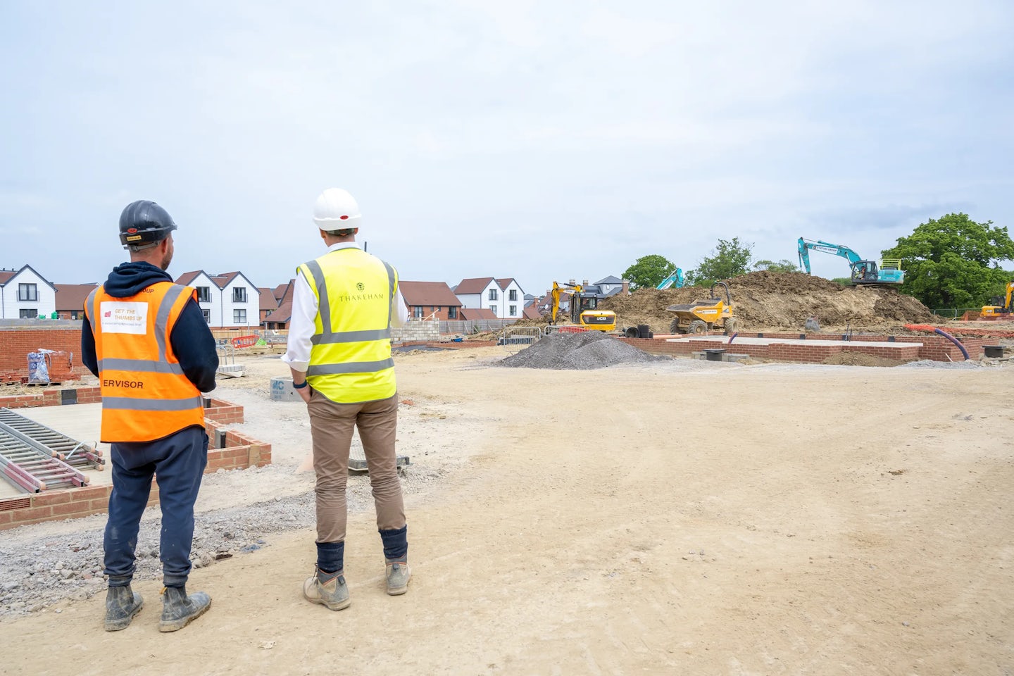 Two construction workers stand on a building site. One wears a yellow high-visibility vest labeled "SUPERVISOR" and a black helmet, while the other wears a white helmet and a yellow vest with "THAKEHAM" on the back. The site is in early development, with excavators, a bulldozer, and partially built structures in the background. Piles of dirt and materials are scattered around, illustrating the active construction process.