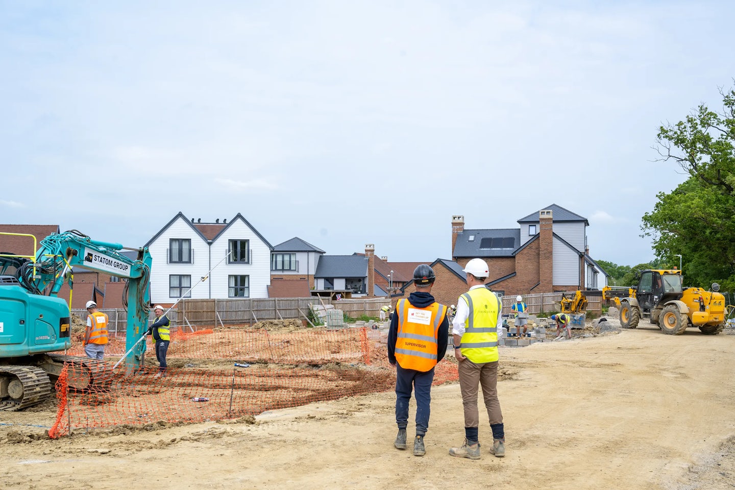 A construction site with workers and heavy machinery. In the foreground, two individuals wearing high-visibility vests and hard hats observe the scene. To the left, a green excavator is in operation, while a yellow loader and a roller are visible in the background. Residential buildings surround the site, suggesting a housing development or urban infrastructure project.