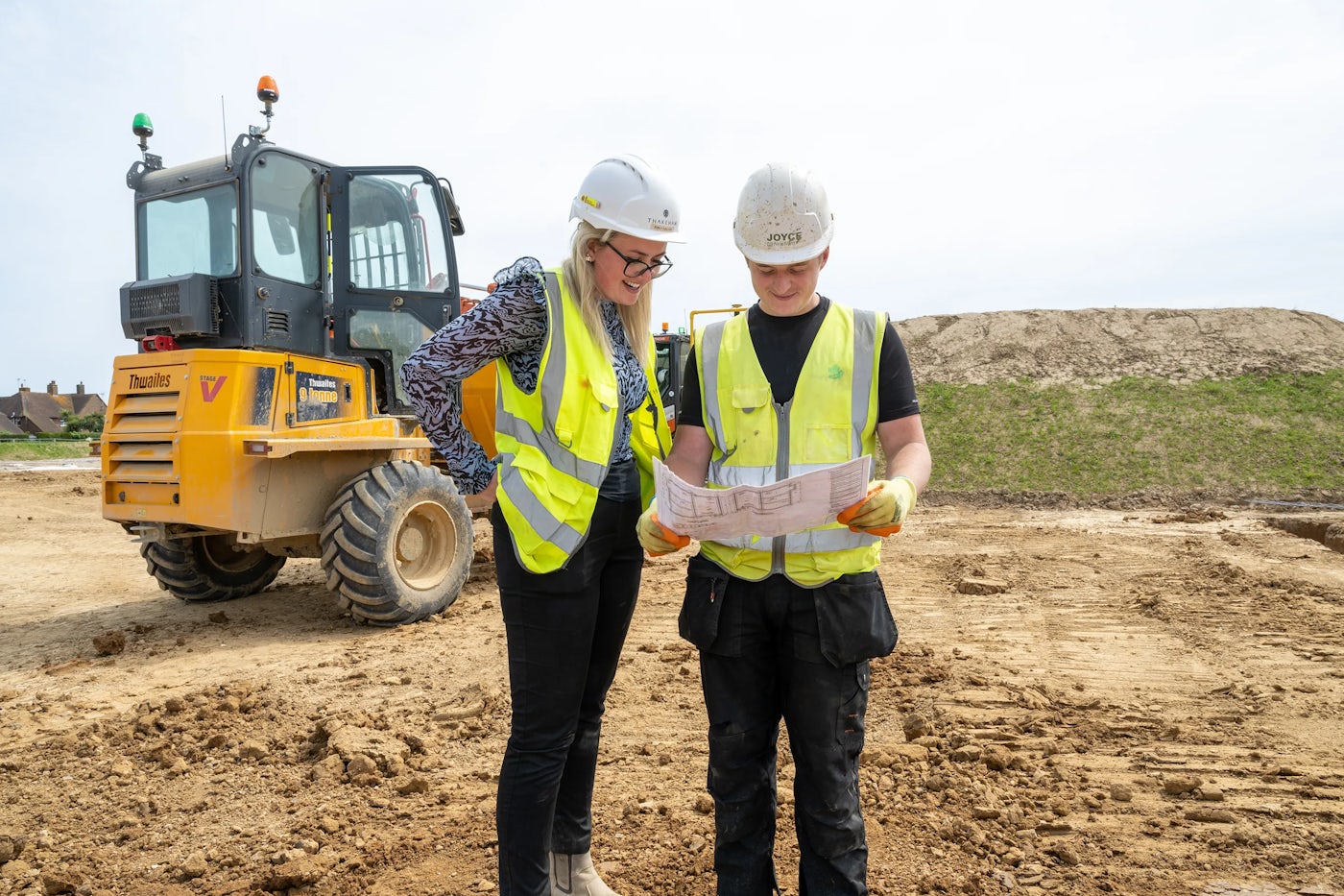 Two construction workers wearing safety helmets and high-visibility vests stand at an active work site. One holds a set of plans, and both examine them intently. Behind them, a yellow dumper truck and a mound of dirt indicate ongoing site activity.