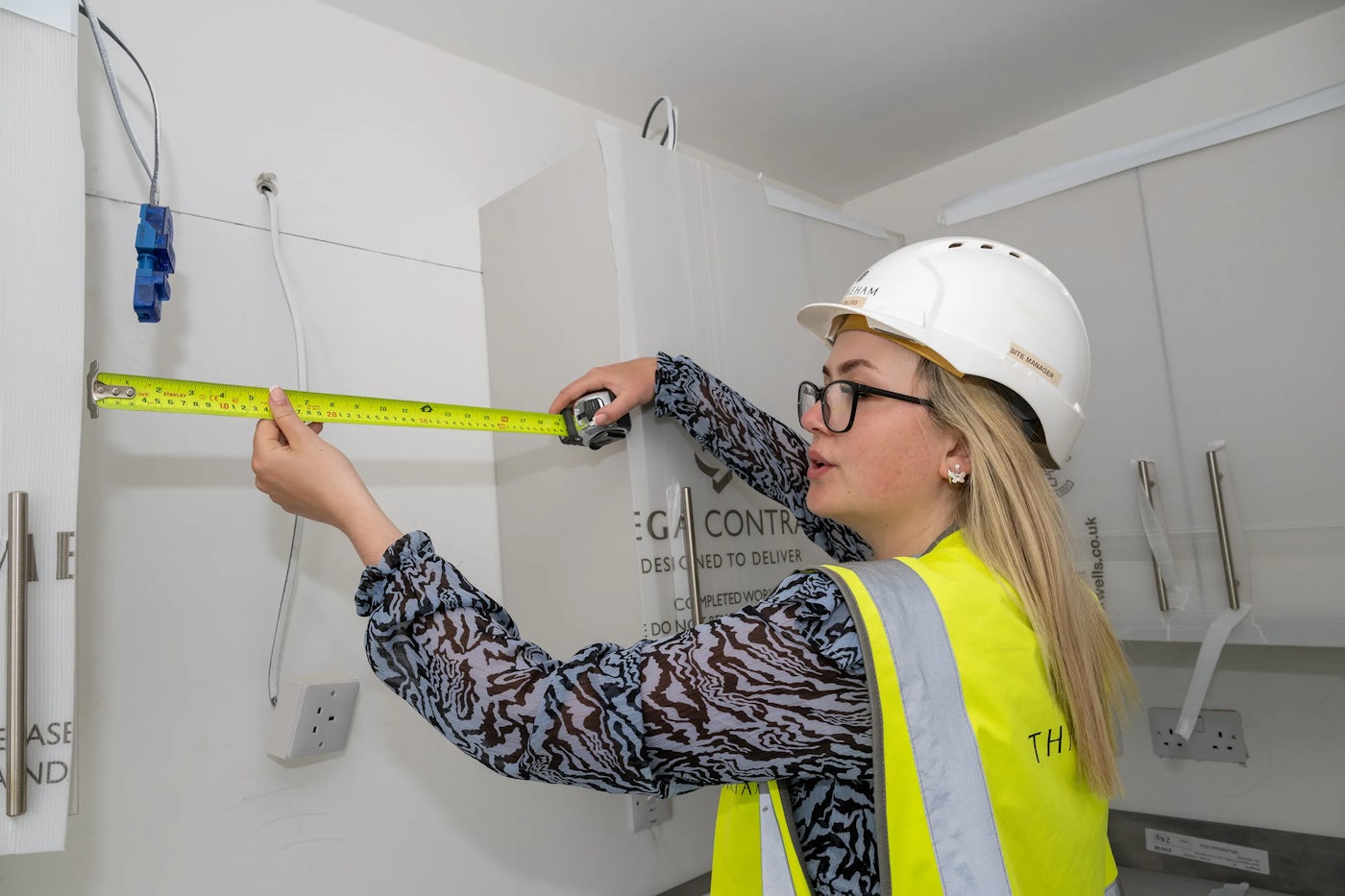 A person wearing a hard hat and a high-visibility vest is using a tape measure to gauge the distance between two points on a wall. Electrical outlets and cables are visible, suggesting the work involves electrical installations or cabinetry. The setting appears to be a construction or renovation site, emphasizing the practical use of measurement in a professional environment.
