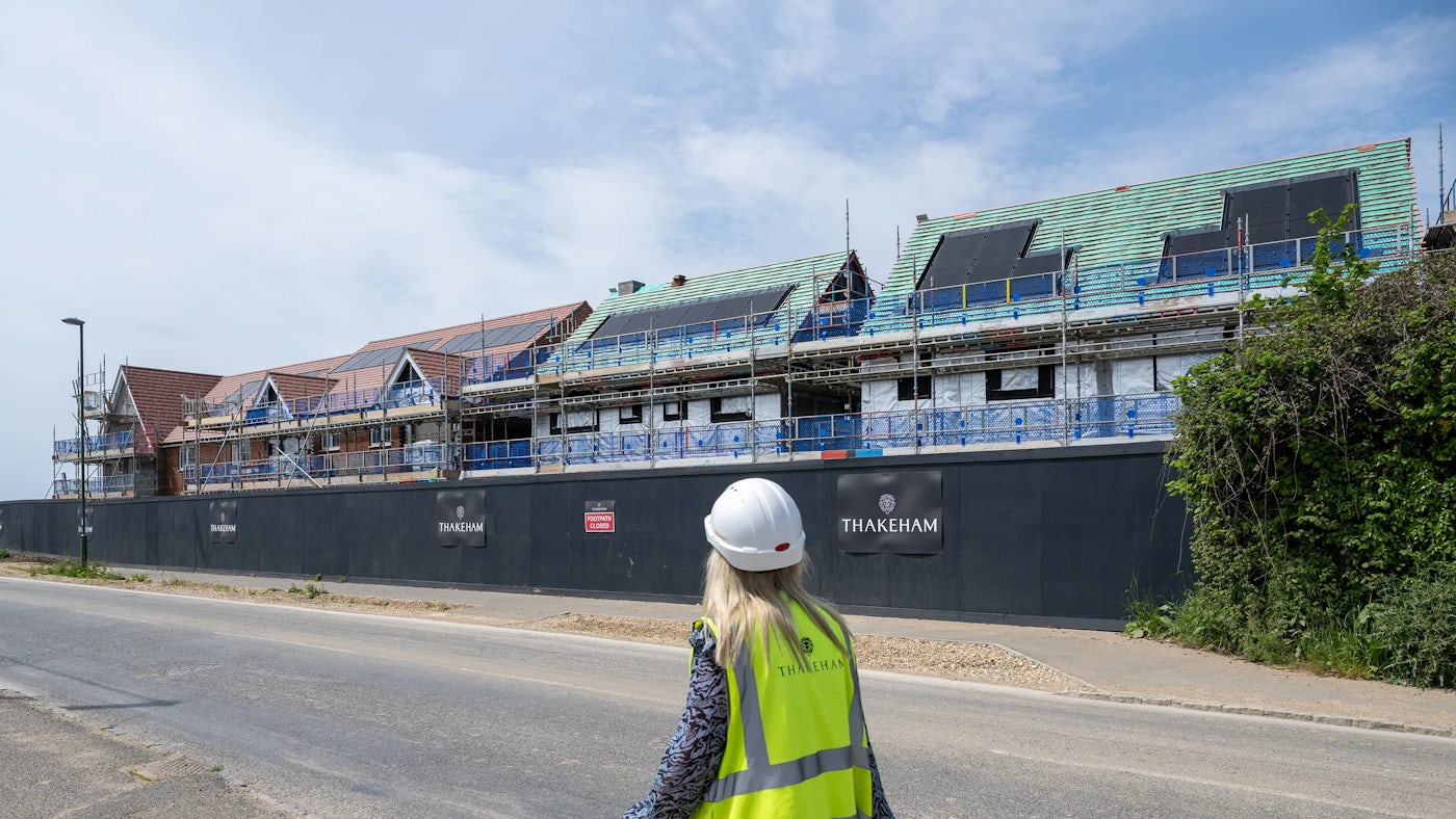 A construction site featuring several houses in various stages of development. Scaffolding surrounds the buildings, with partially tiled roofs in green and red. In the foreground, a person wearing a high-visibility vest and a white hard hat observes the progress. The vest displays the text "THAKEHAM," which matches the branding on the black construction barriers enclosing the site.