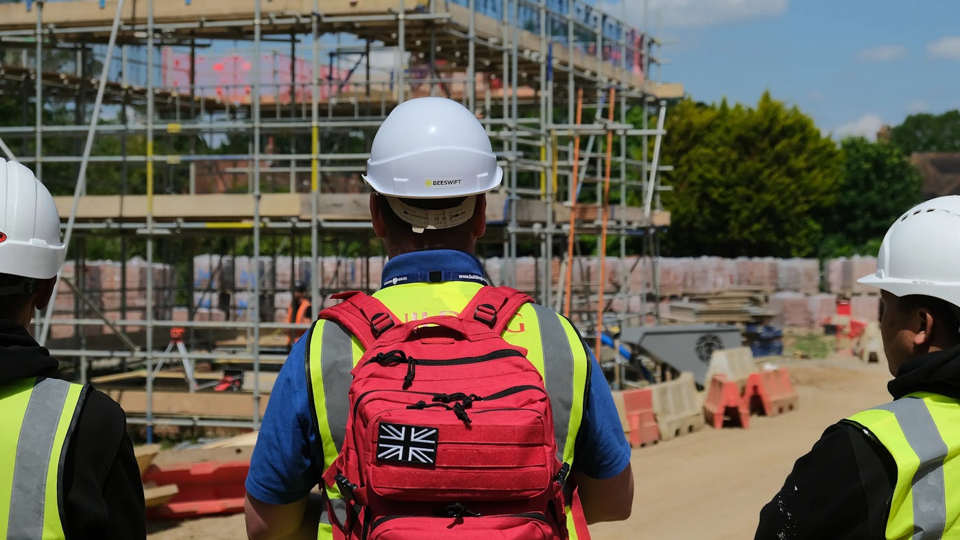 A construction site where three individuals, wearing safety helmets and high-visibility vests, stand together. The person in the center carries a red backpack featuring a Union Jack patch and a black horse emblem. Scaffolding, building materials, and safety barriers are visible in the background, emphasizing the active work environment. The image highlights teamwork and adherence to safety protocols.