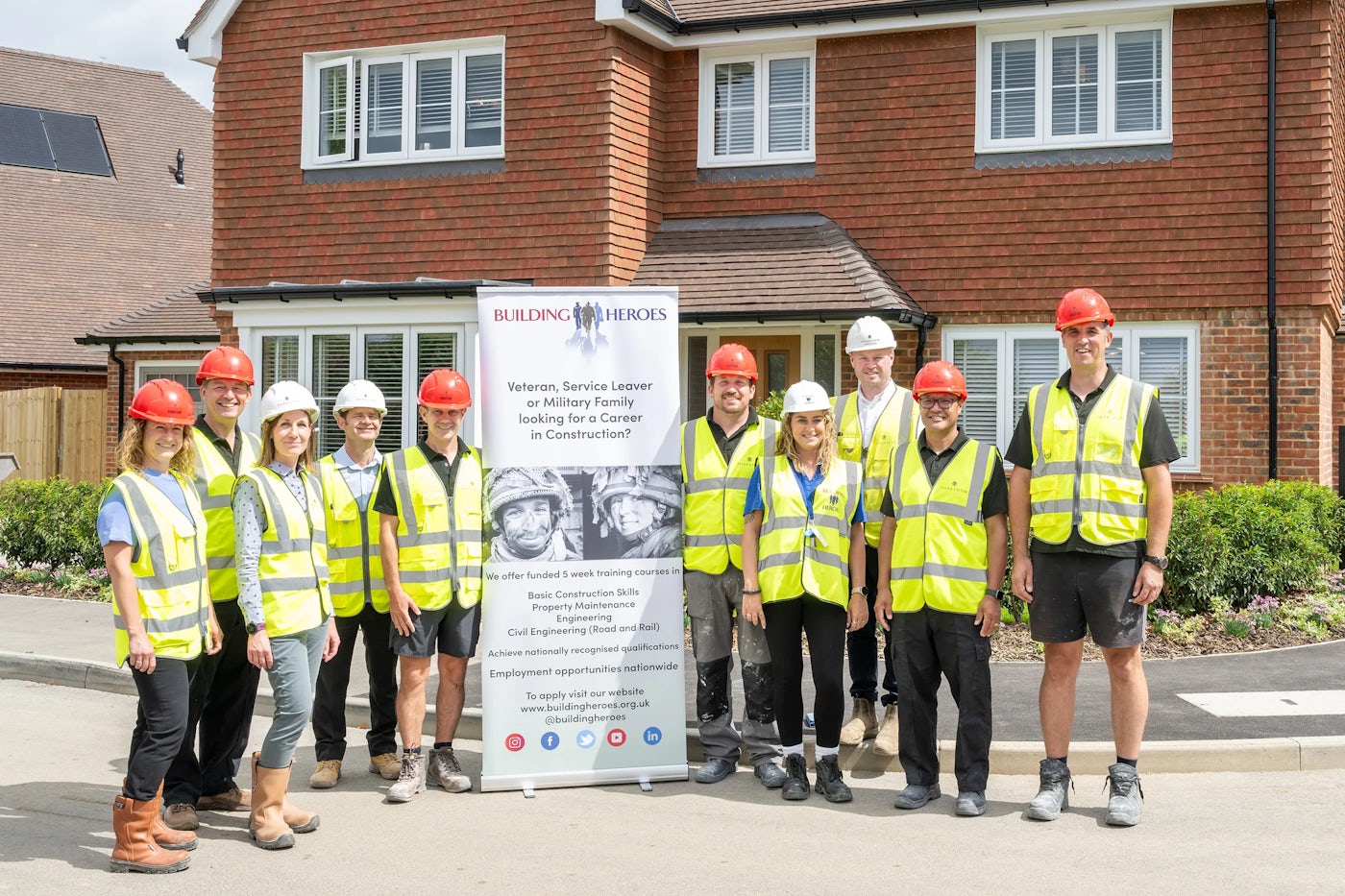 A group of people wearing high-visibility vests and hard hats stand in front of a banner promoting "Building Heroes," an organization supporting veterans, service leavers, and military families seeking construction careers.