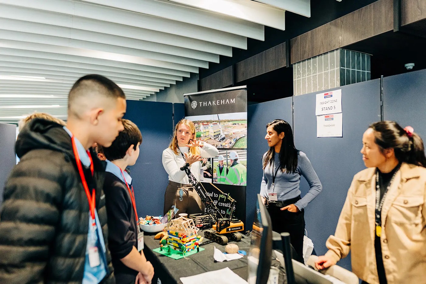 A group of people gathered around a table at a construction-related event or exhibition. The table displays models and equipment, with a banner behind it featuring the name Thakeham and an image of a construction site. A sign on the wall reads Stand 5 and Thakeham, indicating the booth’s location within the event space.