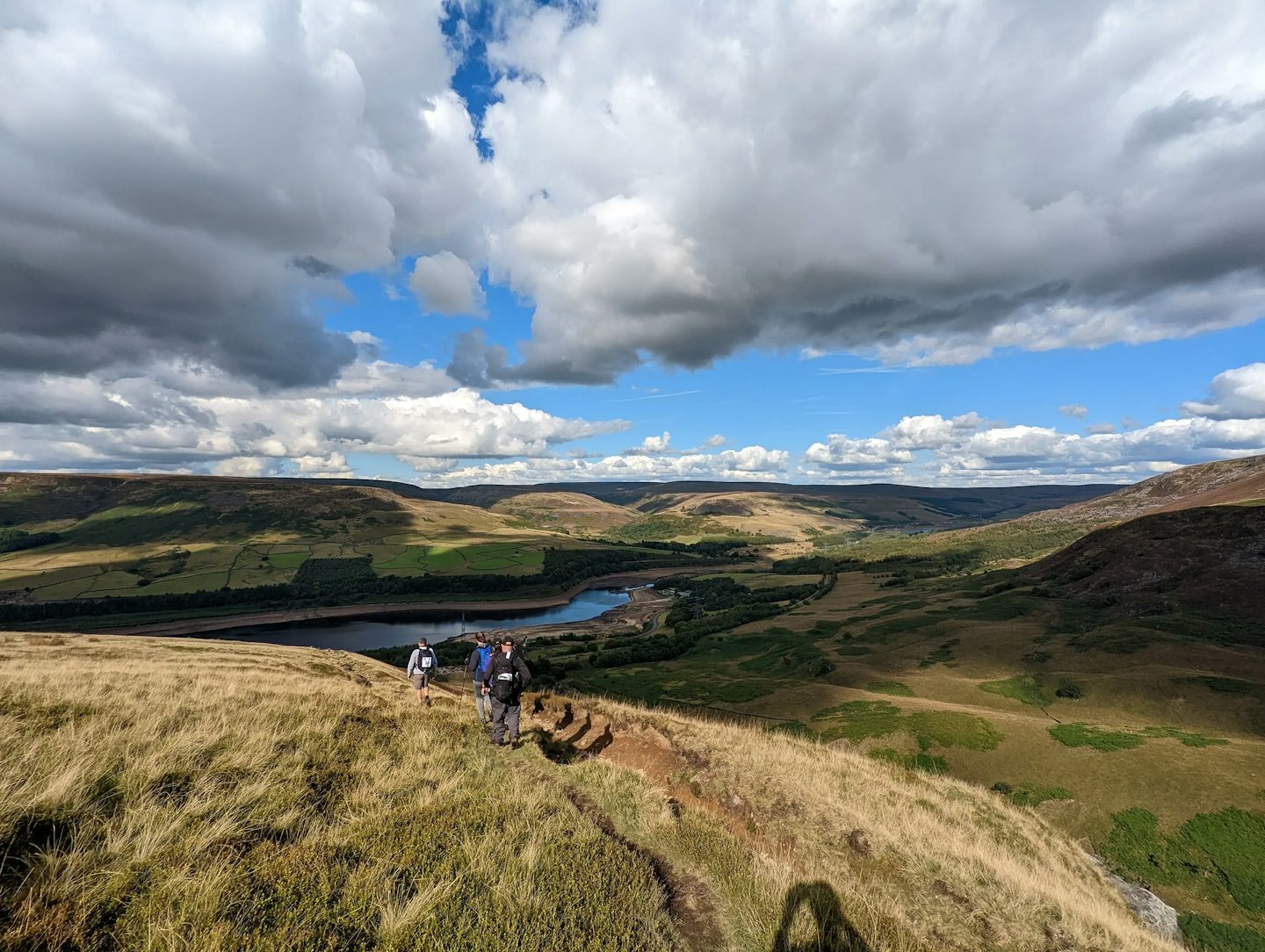 A scenic landscape with a group of hikers walking along a grassy hillside trail. The view features rolling hills, a winding river or reservoir, and a mix of green fields and wooded areas. The sky is partly cloudy with large, fluffy clouds and patches of blue, creating a picturesque outdoor setting that highlights nature and adventure.