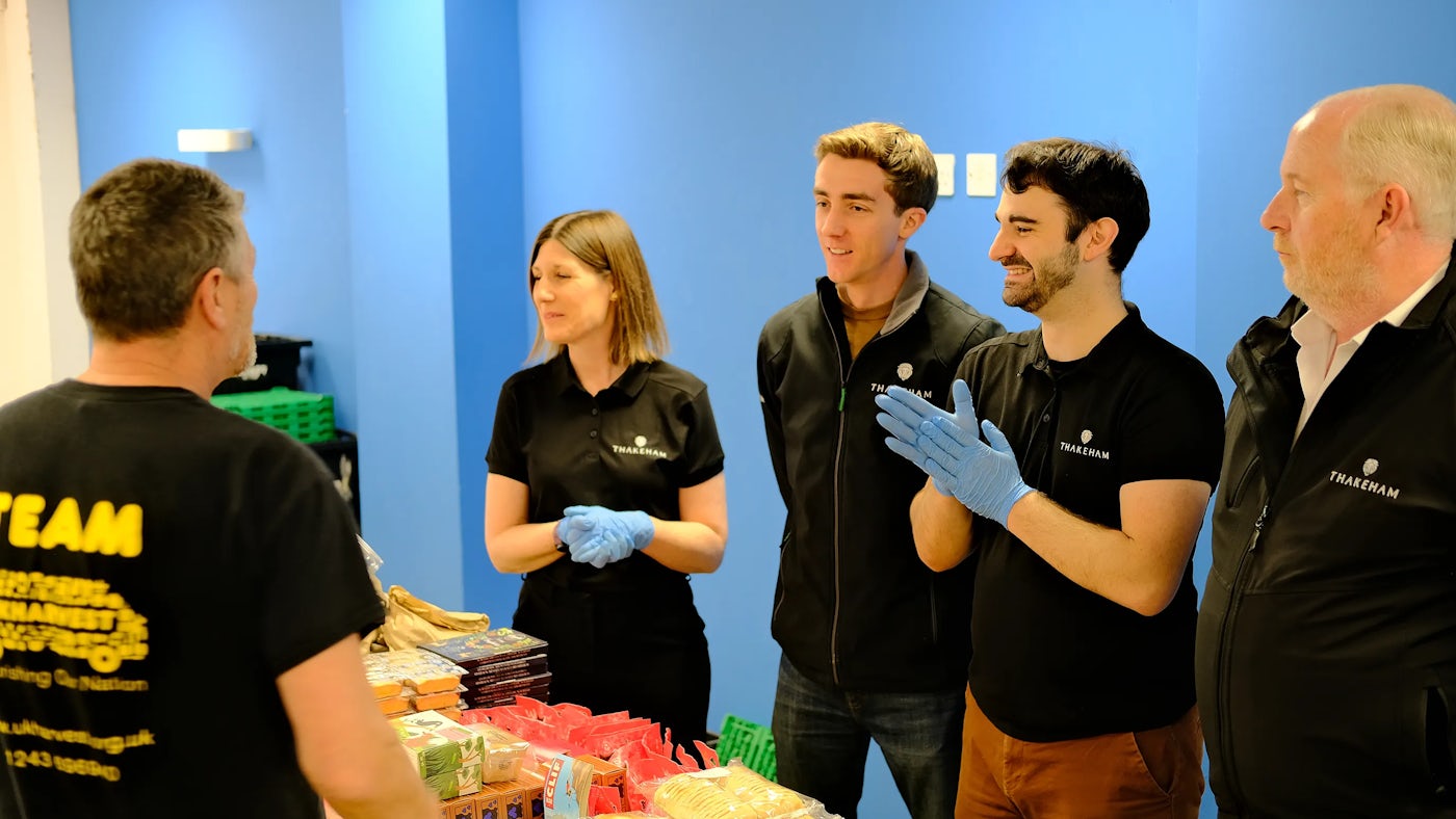 A group of people stand in a room with blue walls, engaged in a discussion around a table filled with various food items, including packaged goods and fresh produce. Four individuals wear black shirts featuring the Thakeham logo.