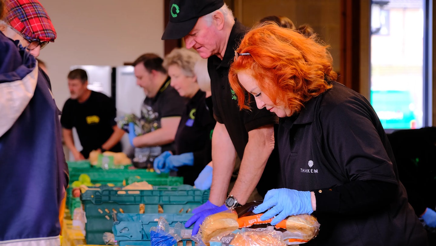 A group of people, wearing black shirts and gloves, are working together at a food distribution event. They are handling various food items, including bread, which is placed in green plastic crates on a table. One person is wearing a black cap.