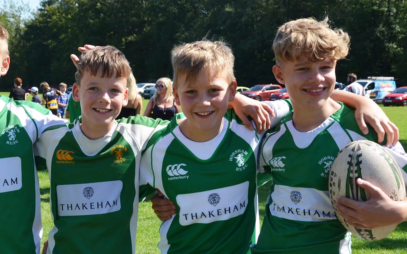 Four young individuals stand on a grassy field, wearing matching green and white rugby jerseys featuring the "Horsham Rugby" team logo and "Thakeham" sponsor branding. They have their arms around each other, displaying camaraderie.