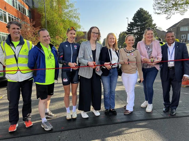 A group of eight people stand in a row outdoors, holding a red ribbon in preparation for a ribbon-cutting ceremony. Some wear casual clothing, while others are dressed in athletic gear or formal attire.
