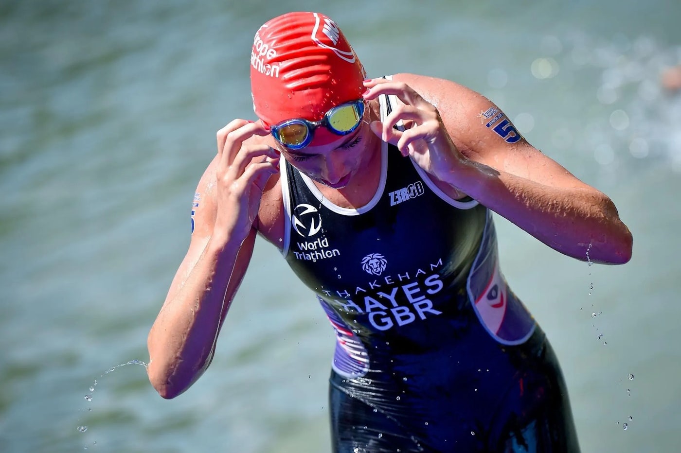 A triathlete emerges from the water during a race, wearing a red swim cap and a black wetsuit adorned with various logos and text, including World Triathlon, THAKEHAM, and HAYES GBR.