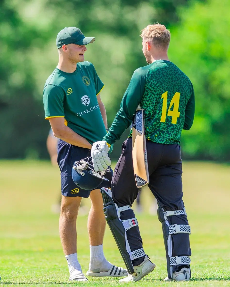 Two cricket players on a field engaged in a discussion. One wears a green and yellow uniform featuring the word "THAKEHAM" and a lion logo on the front, while the other wears a green and black uniform with the number 14 on the back, holding a bat and equipped with protective gear including pads and gloves.