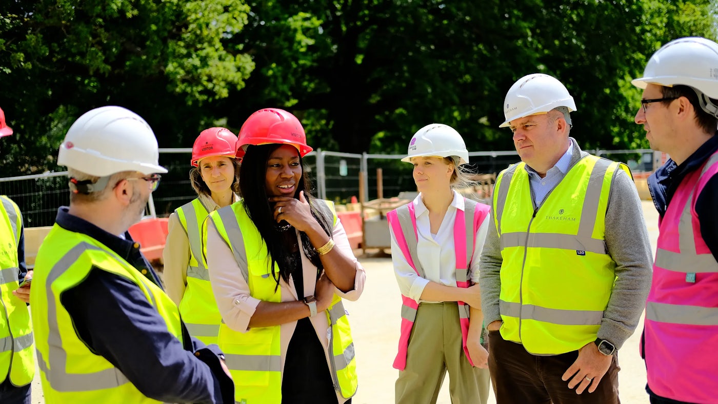 A group of people wearing high-visibility vests and hard hats are gathered at a construction site, engaged in conversation. The background features construction barriers and equipment, reinforcing the active work environment. The individuals appear to be discussing project details, emphasizing collaboration and safety within the construction industry.