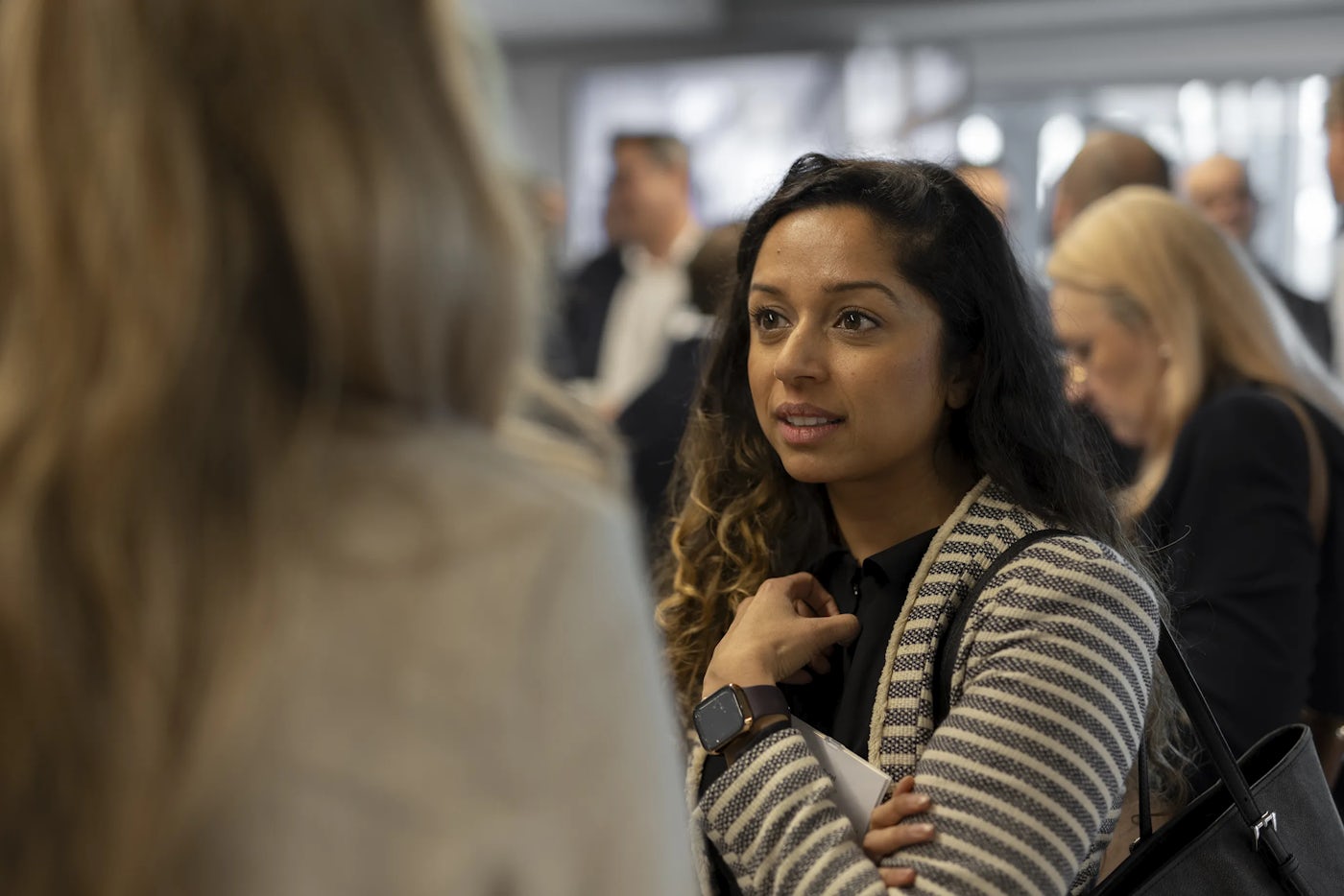 A group of people engaged in conversation at a professional or social gathering. The focus is on an individual wearing a striped sweater and a smartwatch, holding a piece of paper while interacting with another person in the foreground. The blurred background suggests a busy environment with multiple attendees.
