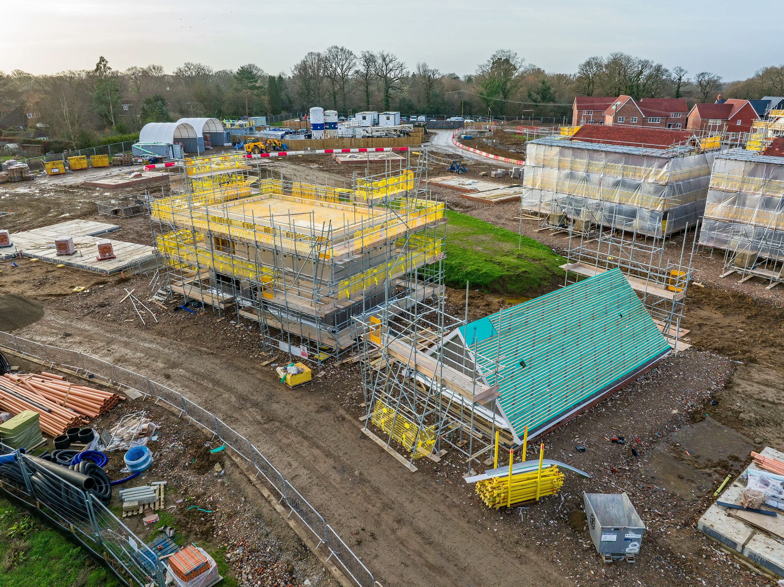 A busy construction site with multiple buildings in various stages of development. A large crane in the center lifts materials to a structure wrapped in yellow scaffolding. Several construction workers, dressed in high-visibility gear and helmets, are dispersed across the site, engaged in various tasks. Heavy machinery and construction vehicles, including excavators and cement mixers, are positioned around the dirt-covered ground, which has patches of green grass.