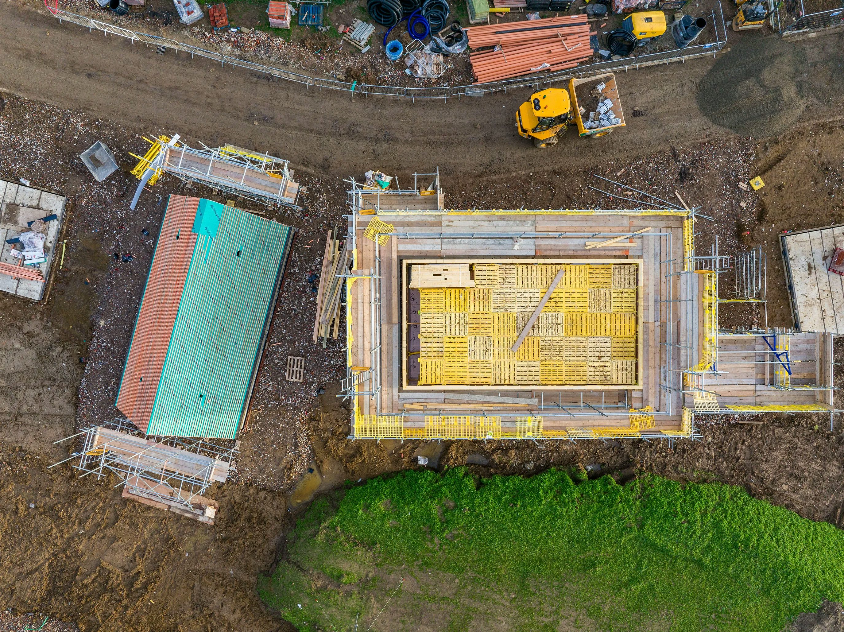 A busy construction site with multiple buildings in various stages of development. A large crane in the center lifts materials to a structure wrapped in yellow scaffolding. Several construction workers, dressed in high-visibility gear and helmets, are dispersed across the site, engaged in various tasks. Heavy machinery and construction vehicles, including excavators and cement mixers, are positioned around the dirt-covered ground, which has patches of green grass.
