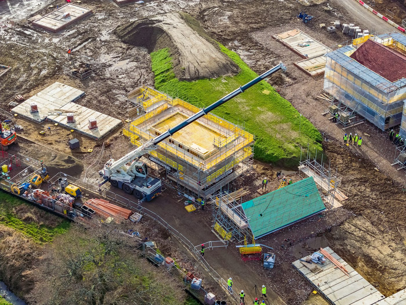 A busy construction site with multiple buildings in various stages of development. A large crane in the center lifts materials to a structure wrapped in yellow scaffolding. Several construction workers, dressed in high-visibility gear and helmets, are dispersed across the site, engaged in various tasks. Heavy machinery and construction vehicles, including excavators and cement mixers, are positioned around the dirt-covered ground, which has patches of green grass.