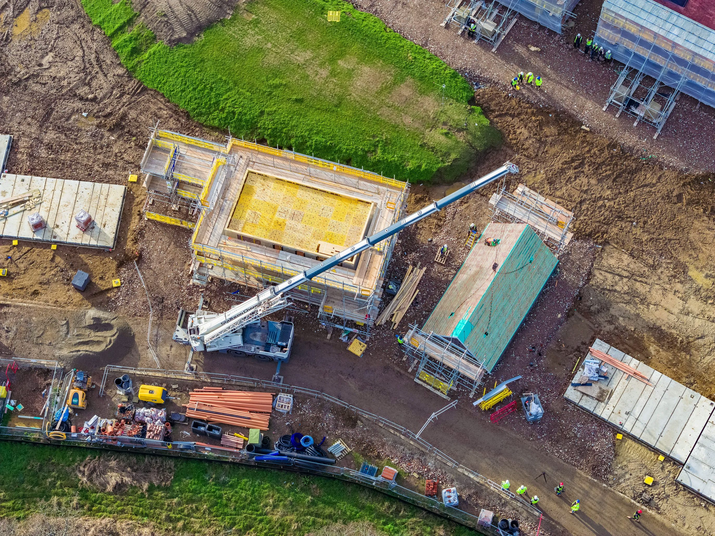 A busy construction site with multiple buildings in various stages of development. A large crane in the center lifts materials to a structure wrapped in yellow scaffolding. Several construction workers, dressed in high-visibility gear and helmets, are dispersed across the site, engaged in various tasks. Heavy machinery and construction vehicles, including excavators and cement mixers, are positioned around the dirt-covered ground, which has patches of green grass.