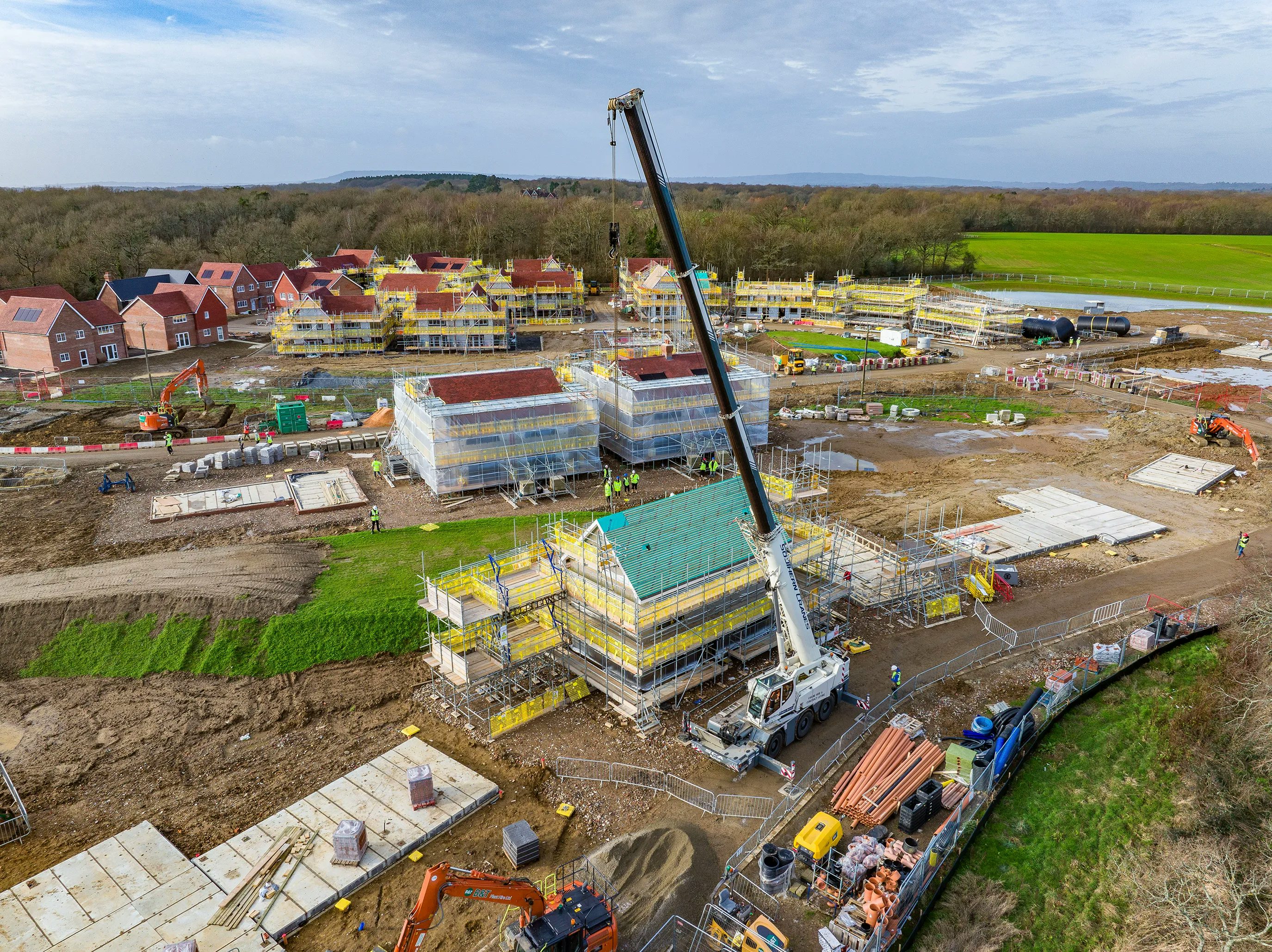 A busy construction site with multiple buildings in various stages of development. A large crane in the center lifts materials to a structure wrapped in yellow scaffolding. Several construction workers, dressed in high-visibility gear and helmets, are dispersed across the site, engaged in various tasks. Heavy machinery and construction vehicles, including excavators and cement mixers, are positioned around the dirt-covered ground, which has patches of green grass.