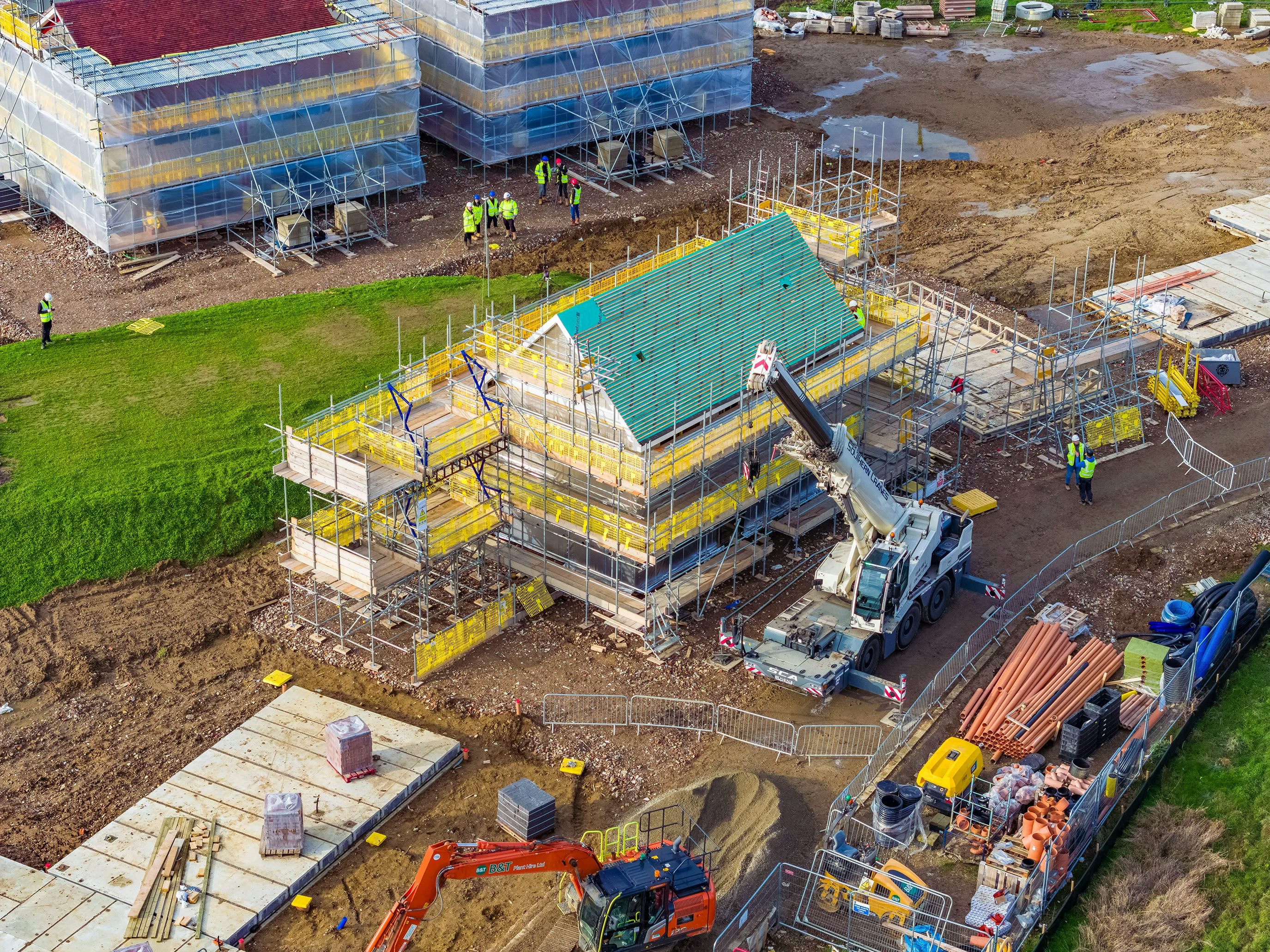 A busy construction site with multiple buildings in various stages of development. A large crane in the center lifts materials to a structure wrapped in yellow scaffolding. Several construction workers, dressed in high-visibility gear and helmets, are dispersed across the site, engaged in various tasks. Heavy machinery and construction vehicles, including excavators and cement mixers, are positioned around the dirt-covered ground, which has patches of green grass.