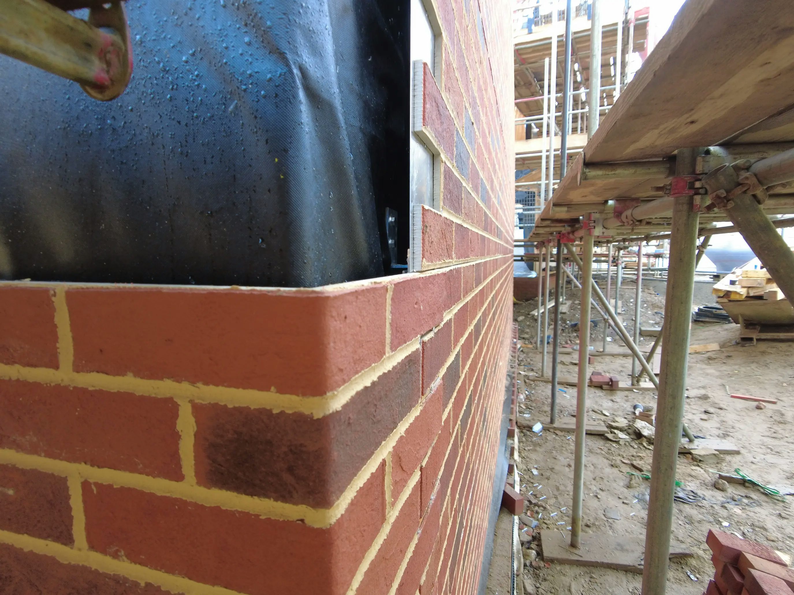 A construction site featuring a partially assembled printed brick wall, designed to resemble traditional masonry with red and dark brown tones arranged in a staggered pattern. Yellow mortar-like material fills the spaces, reinforcing the realistic appearance. A black waterproof membrane is partially visible at the top left, while scaffolding on the right supports the ongoing process. Various construction materials and debris lie scattered on the ground, emphasizing the precision and efficiency of modern building techniques.