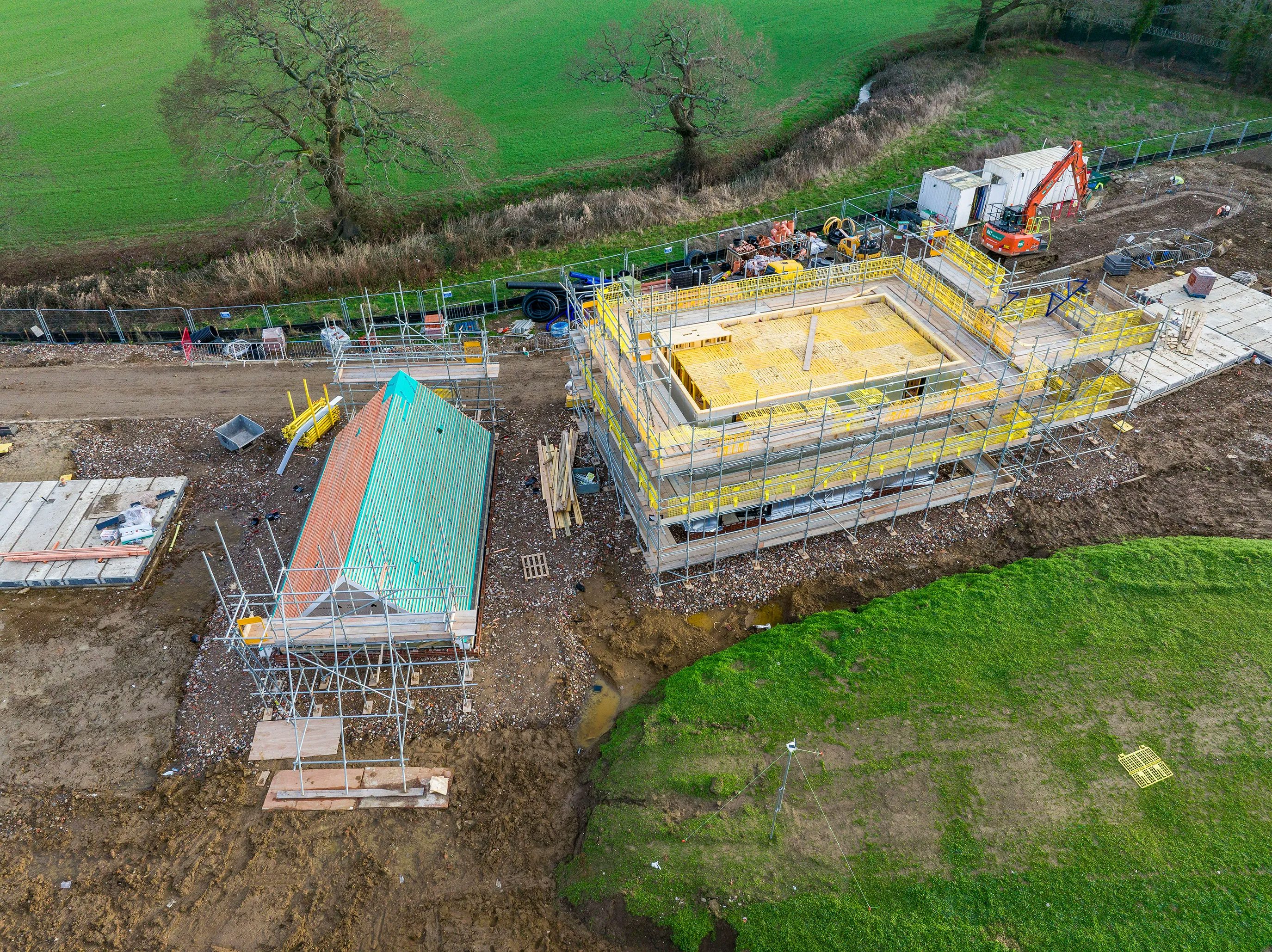 A construction site with various active projects and equipment. In the foreground, a yellow truck is parked near stacks of wooden planks and metal pipes. A partially built structure with scaffolding surrounds its exterior, and a green roof is visible on another nearby building.