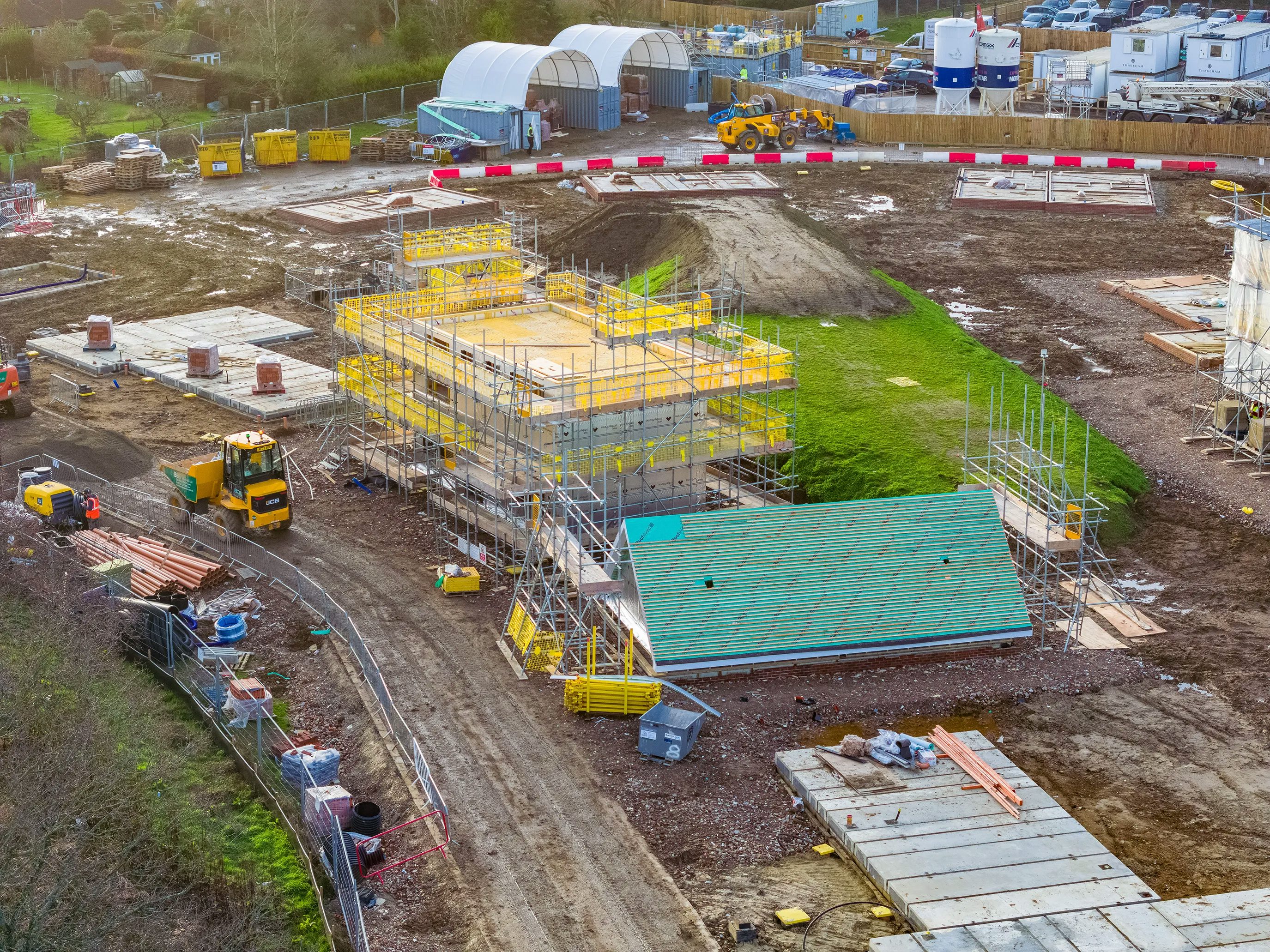 A construction site with various active projects and equipment. In the foreground, a yellow truck is parked near stacks of wooden planks and metal pipes. A partially built structure with scaffolding surrounds its exterior, and a green roof is visible on another nearby building.