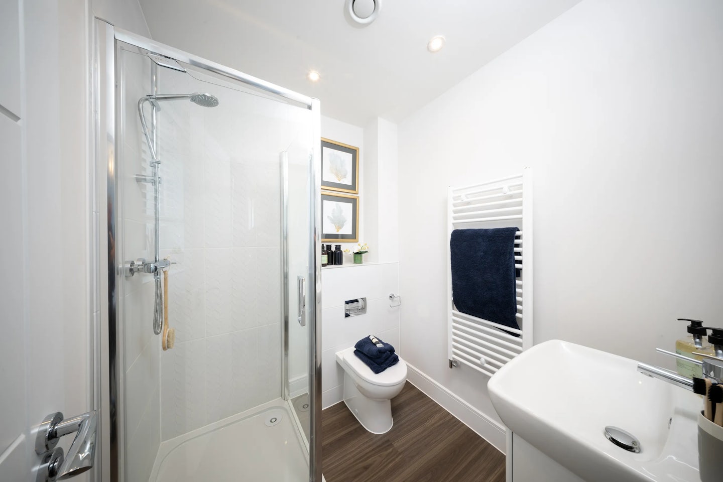 A modern bathroom featuring a glass-enclosed shower with a chrome showerhead and handle on the left. Next to the shower, a toilet sits beneath a small shelf holding toiletries and a potted plant, with a folded dark blue towel placed on top. Three framed pictures decorate the wall above the shelf. On the right, a white towel radiator holds a matching dark blue towel, and a sleek white sink with a chrome faucet is accompanied by neatly arranged toiletries. The room has white walls and a wooden floor, creating a clean, contemporary aesthetic, illuminated by recessed ceiling lights.