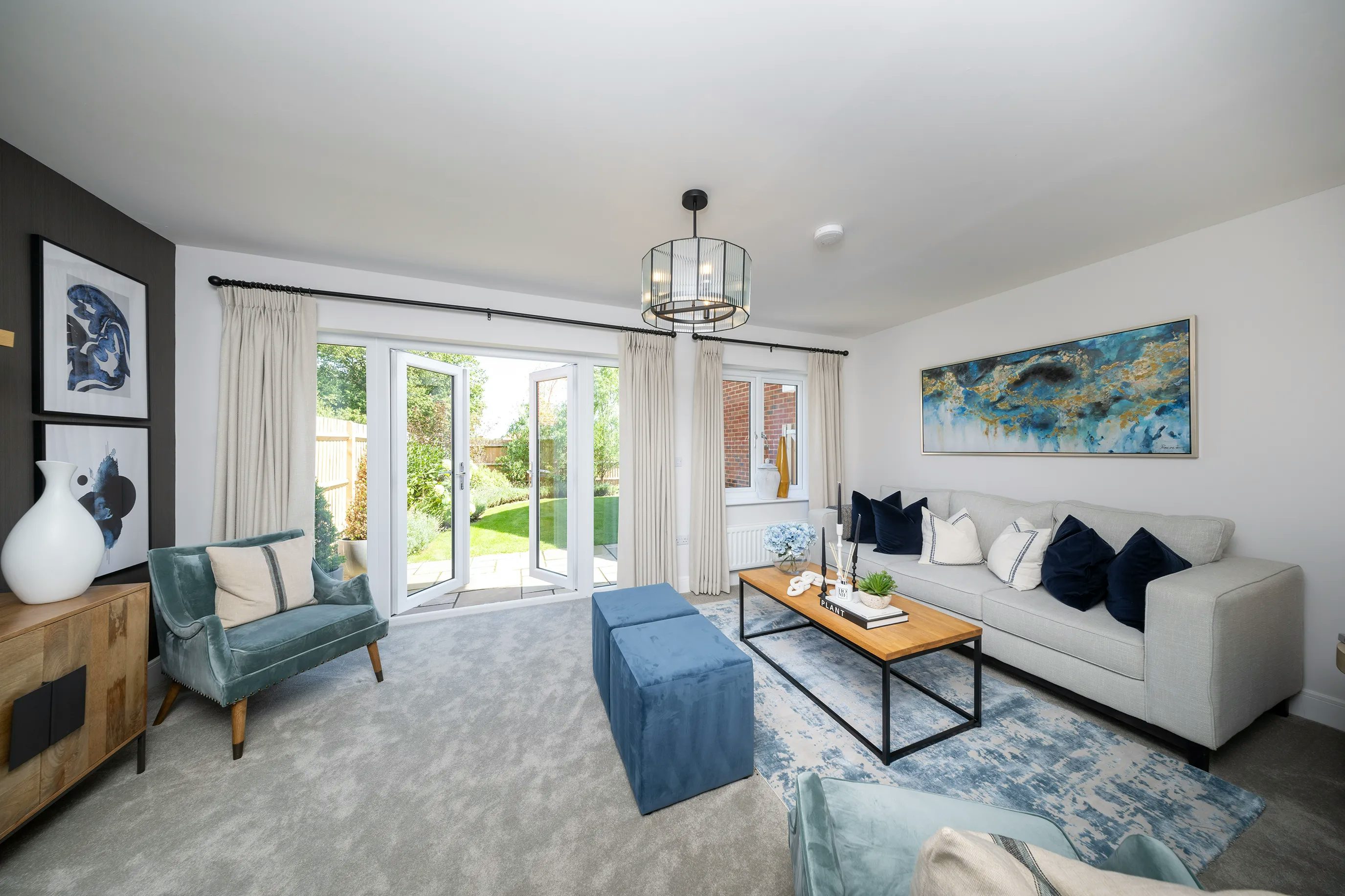 A bright and elegant modern living room with a spacious layout. The room features a large gray sofa adorned with white and navy blue pillows, complemented by a wooden coffee table with decorative items. A blue patterned rug adds texture beneath two blue upholstered chairs, one draped with a white throw blanket. Against one wall, a wooden sideboard holds decorative vases, with framed artwork displayed above. Large glass doors open to a garden, allowing abundant natural light to enhance the room’s airy feel. A contemporary chandelier hangs from the ceiling, while a large abstract painting above the sofa adds a stylish touch.