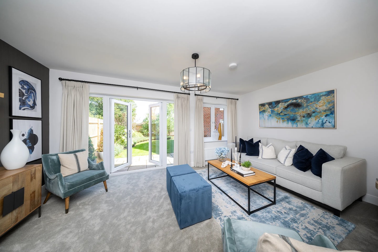 A bright and elegant modern living room with a spacious layout. The room features a large gray sofa adorned with white and navy blue pillows, complemented by a wooden coffee table with decorative items. A blue patterned rug adds texture beneath two blue upholstered chairs, one draped with a white throw blanket. Against one wall, a wooden sideboard holds decorative vases, with framed artwork displayed above. Large glass doors open to a garden, allowing abundant natural light to enhance the room’s airy feel. A contemporary chandelier hangs from the ceiling, while a large abstract painting above the sofa adds a stylish touch.