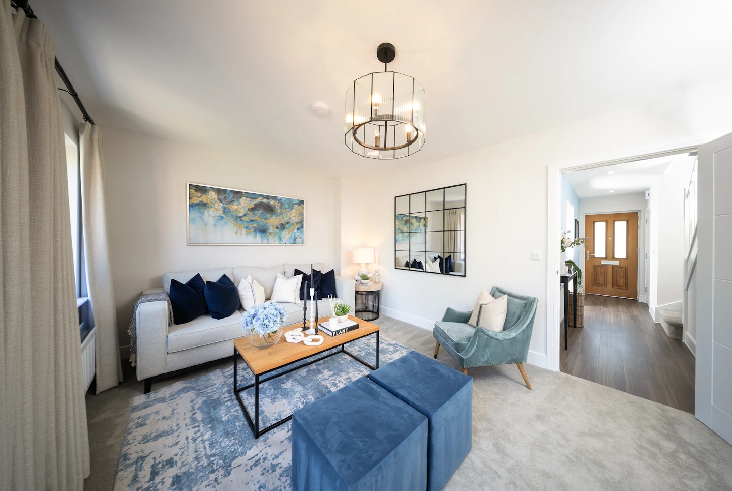 A contemporary living room featuring a white sofa with dark blue cushions, a wooden coffee table with metal legs, and two matching blue ottomans. A light blue armchair with a white pillow sits nearby. The coffee table holds decorative items, including a vase with blue flowers, a small plant, and books. Behind the sofa, a large abstract painting in blue and gold hues hangs next to a grid-patterned mirror. Natural light streams in through the windows, illuminating a stylish chandelier overhead. The floor is covered with a light-colored carpet, with a blue and white patterned rug beneath the coffee table. The room leads into a hallway with wooden flooring and a wooden door at the end.